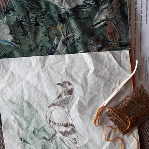 May include: A white fabric panel with a green and brown bird design. The fabric is folded in half and there is a small bag of dried herbs and a length of orange thread on top of the fabric.