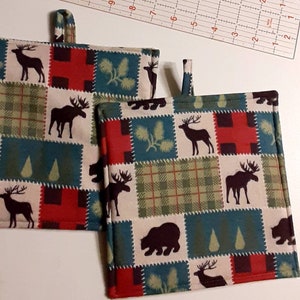 May include: Two red, green, and white patterned pot holders with a woodland theme. The pot holders feature moose, deer, bears, and pine trees.