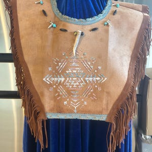 May include: A tan suede vest with a geometric design in turquoise, white, and silver. The vest has brown fringe along the bottom and a turquoise beaded collar. A small antler is attached to the front.