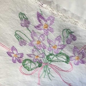 以下が含まれることがあります： ピンクのリボンで結ばれた、紫と黄色の花束が刺繍された白いテーブルクロスです。