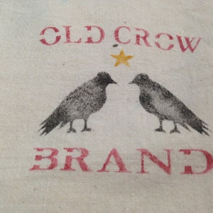 Może przedstawiać: Biały materiał z nadrukiem "Old Crow Brand" w kolorze czerwonym. Dwa czarne ptaki patrzą na siebie, między nimi żółta gwiazda.