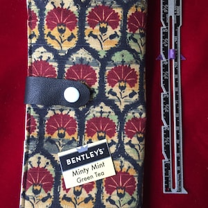 May include: A black fabric wallet with a floral pattern in shades of red, yellow, and green. The wallet has a black leather strap with a white button. The wallet has a label that reads "BENTLEY'S Minty Mint Green Tea".