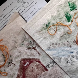 May include: Two pieces of white fabric with brown, red, and green paint splatters. Each piece has a red house shape printed on it. A yellow rubber band is on each piece of fabric.