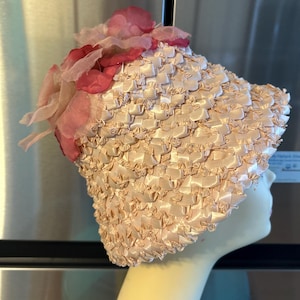 May include: A vintage-style hat in a light peach color, featuring a woven texture and adorned with pink fabric flowers. The hat is displayed on a mannequin head, showcasing its design and shape. The hat's texture appears to be made of woven ribbons.
