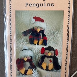 May include: A Homespun Penguins pattern featuring three handmade penguin dolls. Each penguin is approximately 8 inches tall and wears a hat, scarf, or earmuffs. The penguins have yellow feet and beaks, with black and red accents.