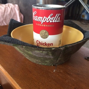 May include: A red and white can of Campbell's Condensed Chicken Noodle Soup sitting inside a camouflage baseball cap.