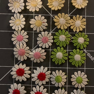 May include: Assortment of 18 white daisy-shaped fabric appliques with various colored centers. The centers include pink, red, green, and yellow. The appliques are arranged in rows on a black and white grid.