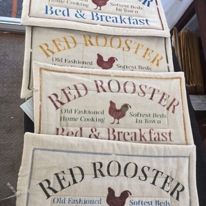 May include: Four stacked fabric signs with the text "RED ROOSTER" in a red, yellow, and blue font. Each sign has a red rooster illustration and the text "Old Fashioned Home Cooking" and "Softest Beds In Town" and "Bed & Breakfast".