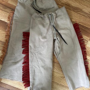 May include: Two-piece beige outfit with drawstring waist and red fringe accents. The outfit includes a skirt and pants, both made of a light-colored fabric. The skirt has fringe along the side, and the pants have fringe along the outer leg.