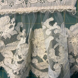 May include: An off-white lace trim featuring a scalloped edge with intricate floral embroidery and a sheer tulle base. The top section is finished with a decorative braided gimp trim adorned with small, evenly spaced white beads.