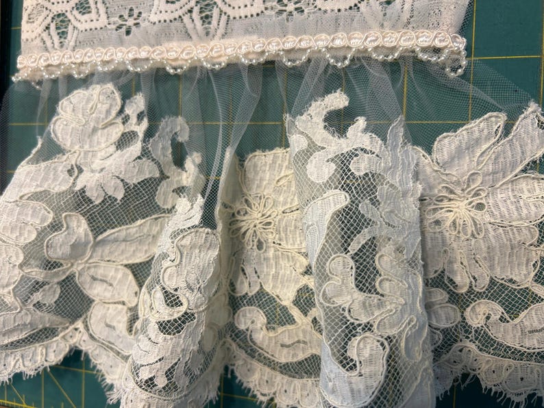 May include: An off-white lace trim featuring a scalloped edge with intricate floral embroidery and a sheer tulle base. The top section is finished with a decorative braided gimp trim adorned with small, evenly spaced white beads.