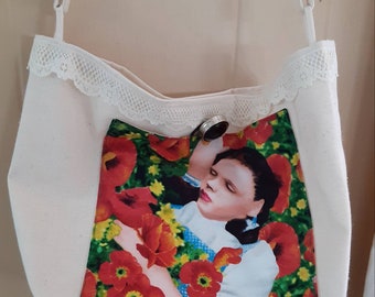 Puede incluir: Una bolsa de lona blanca con un borde de encaje y una imagen impresa de una niña con un vestido azul y blanco tumbada en un campo de amapolas rojas. La bolsa tiene un anillo plateado sujeto a la correa.