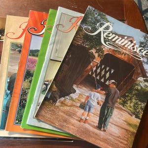 Könnte beinhalten: Ein Stapel Vintage-Magazine "Reminisce" mit farbenfrohen Covern und nostalgischen Bildern. Das oberste Magazin zeigt ein Gemälde einer überdachten Brücke und zwei Personen beim Angeln. Andere Cover zeigen Szenen aus Natur und häuslichem Leben.