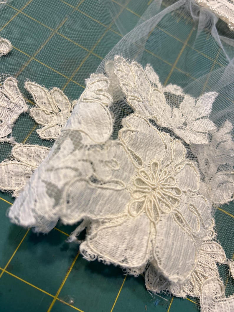 May include: An off-white lace applique featuring intricate floral and leaf patterns attached to a sheer, fine-mesh tulle fabric. The lace design displays detailed embroidery with scalloped edges and delicate thread work throughout the textile.