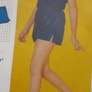 Può includere: Una ragazza giovane che indossa un vestito blu senza maniche con bordi bianchi e scarpe rosse. Il vestito ha un nodo di corda bianco sul collo. La ragazza è in piedi su uno sfondo giallo.