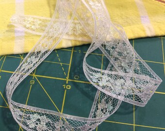 Puede incluir: Una cinta de encaje blanco transparente con un patrón floral y acentos brillantes. La cinta está rizada y descansa sobre una superficie amarilla y verde, lo que sugiere un proyecto de artesanía o costura.