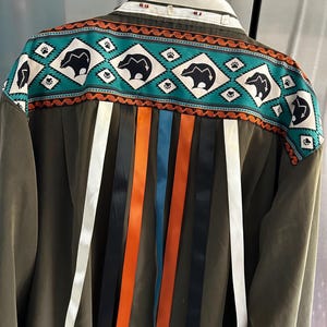May include: Olive green corduroy shirt with a decorative yoke featuring bear silhouettes, turquoise, and orange geometric patterns. Colorful ribbons in white, black, orange, and blue hang from the back, adding a unique touch to this garment.