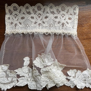 May include: An off-white lace trim featuring a wide, intricate floral pattern at the top edge and a sheer, lightweight tulle section below. The bottom border is finished with large, detailed floral lace appliqu&eacute;s in a matching off-white shade.