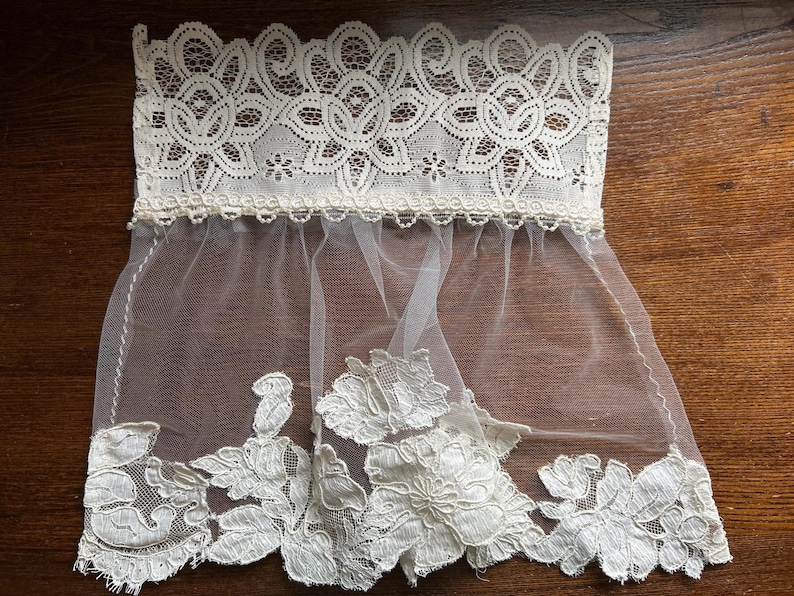 May include: An off-white lace trim featuring a wide, intricate floral pattern at the top edge and a sheer, lightweight tulle section below. The bottom border is finished with large, detailed floral lace appliqu&eacute;s in a matching off-white shade.