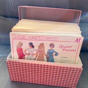 May include: A red and white gingham patterned box filled with vintage sewing patterns. The top pattern reads "Classic Waisted Dresses" with illustrations of dresses. The brand name "Creative Patterns" is also visible.