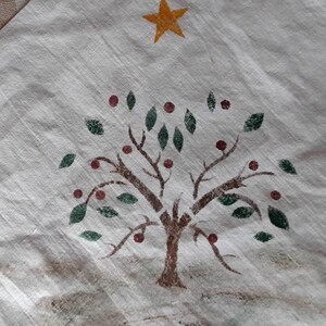 May include: A white fabric square with a brown tree painted on it. The tree has green leaves and red berries. A yellow star is painted at the top of the fabric.