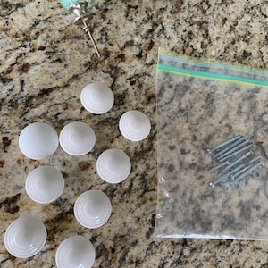 May include: A set of ten white ceramic drawer knobs with a raised, concentric circle design. The knobs are arranged in a semi-circle on a light brown countertop. A clear green glass knob with a silver base is also visible. A plastic bag containing screws is in the background.