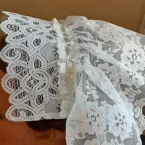 Puede incluir: Un gorro de encaje blanco con un borde festoneado y un lazo de cinta de sat&eacute;n. El gorro est&aacute; hecho de encaje delicado con un patr&oacute;n floral.