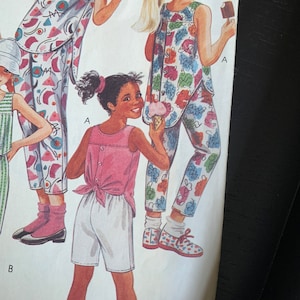 May include: A sewing pattern for children's clothing. The pattern includes a sleeveless top with a tie at the back and shorts. The top has a floral print and the shorts are white. The pattern is illustrated with three children wearing the clothing.