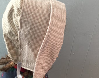 Op de afbeelding: Een beige gingham-bonnet met een wit lint dat onder de kin is gebonden. De bonnet is gemaakt van een zachte, gewatteerde stof en heeft een eenvoudig, klassiek ontwerp.