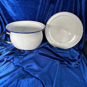 May include: White enamel chamber pot with a blue rim and a black handle, next to a matching lid. The chamber pot is round with a wide opening. The lid is also white and round. The items are on a blue velvet surface.