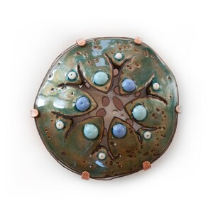 May include: A handcrafted ceramic art piece with a circular design. The piece features a mottled green and brown glaze, accented with light blue and white spherical elements. Copper-colored metal pieces are attached around the edge. The artwork has a unique, organic aesthetic.