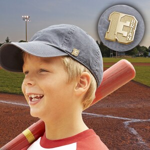 K&ouml;nnte beinhalten: Ein junger Junge tr&auml;gt eine graue Baseballkappe mit einer goldenen Baseballnadel an der Seite. Er h&auml;lt einen Holzbaseballschl&auml;ger und l&auml;chelt. Die Nadel tr&auml;gt die Nummer 13 und den Namen "Brian".