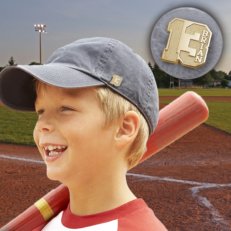 K&ouml;nnte beinhalten: Ein junger Junge tr&auml;gt eine graue Baseballkappe mit einer goldenen Baseballnadel an der Seite. Er h&auml;lt einen Holzbaseballschl&auml;ger und l&auml;chelt. Die Nadel tr&auml;gt die Nummer 13 und den Namen "Brian".