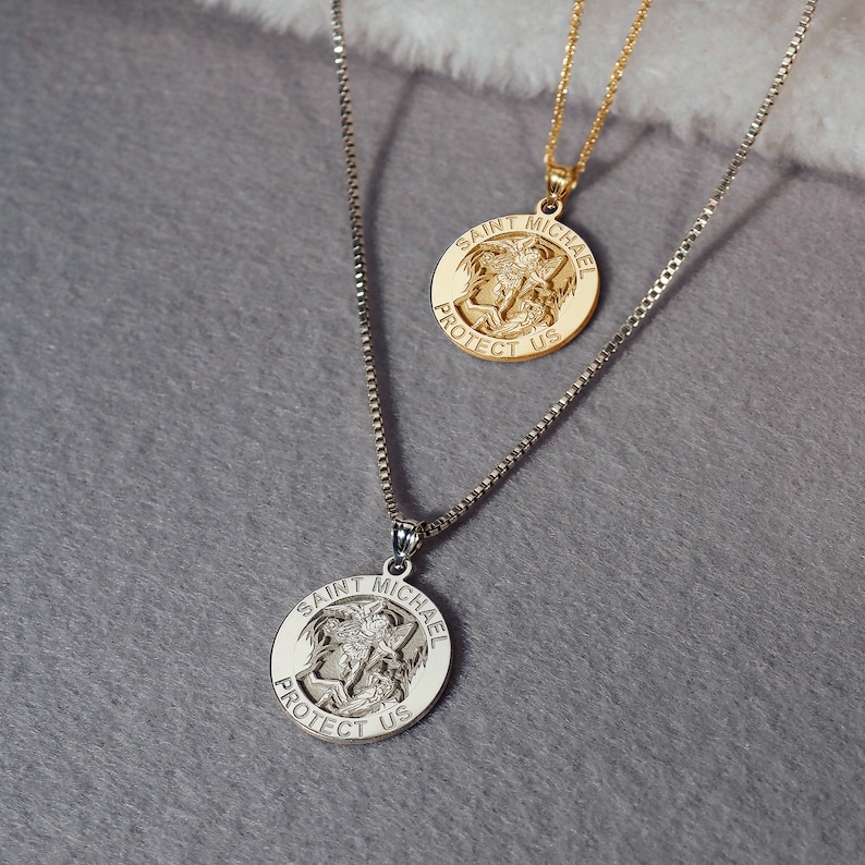 May include: Two gold necklaces with Saint Michael pendants. The pendants feature a depiction of Saint Michael slaying a demon, with the words "Saint Michael Protect Us" inscribed around the edge.