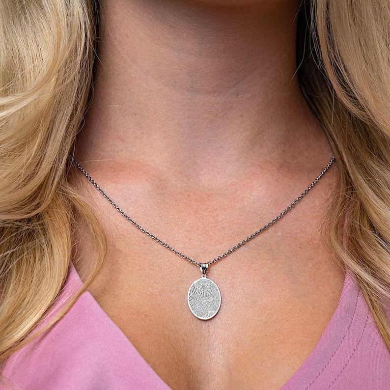 Puede incluir: Un collar de cadena de plata con un colgante redondo que presenta un dise&ntilde;o de huella digital detallado.