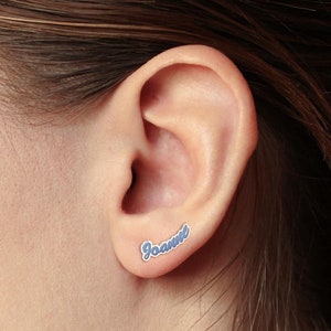 May include: A close-up of a person's ear with a blue and white nameplate earring that says "Joanne".