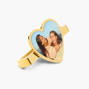 Pu&ograve; includere: Anello dorato a forma di cuore con una foto di due donne che ridono. L'anello &egrave; una semplice fascia con una lunetta a forma di cuore.