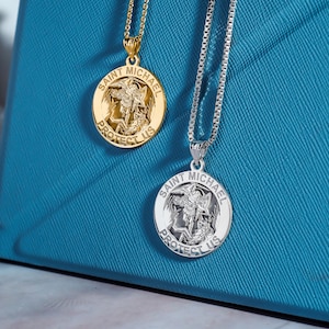May include: Two silver and gold necklaces with Saint Michael medallions. The medallions feature a depiction of Saint Michael slaying a dragon with the words "Saint Michael Protect Us" around the edge.