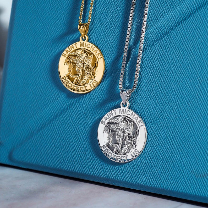 May include: Two silver and gold necklaces with Saint Michael medallions. The medallions feature a depiction of Saint Michael slaying a dragon with the words "Saint Michael Protect Us" around the edge.