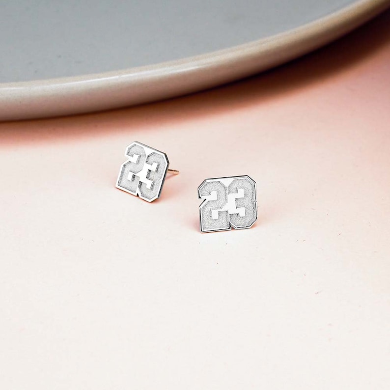 May include: A pair of silver earrings featuring the number 23 in a bold, white font.