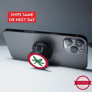 May include: A black smartphone with a pop-up phone grip attached. The grip features a red and white design with a green leaf. The phone grip is attached to the back of the phone. The text "SHIPS SAME OR NEXT DAY" is in a red box.