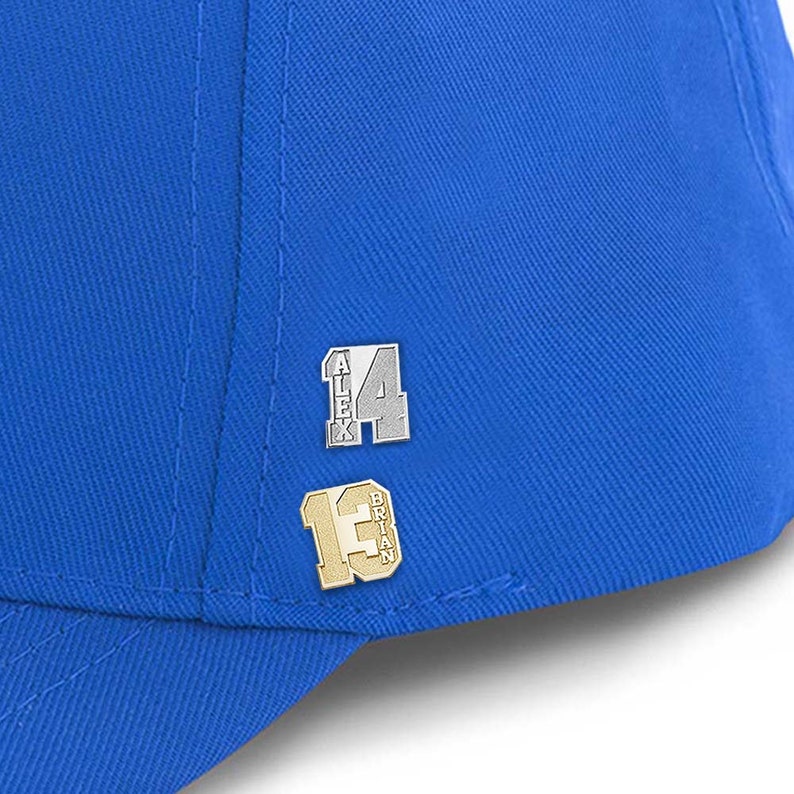 K&ouml;nnte beinhalten: Eine blaue Baseballkappe mit zwei silbernen und goldenen Pins. Der obere Pin sagt "Alex 14" und der untere Pin sagt "Brian 13".