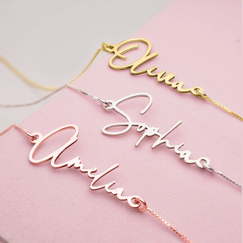 Peut inclure: Trois colliers d&eacute;licats avec des noms personnalis&eacute;s, "Olivia", "Sophia" et "Amelia", &eacute;crits en cursive. Les colliers sont sur un fond rose.
