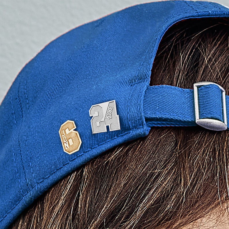 K&ouml;nnte beinhalten: Eine blaue Baseballkappe mit einer goldenen Nummer 6 und einer silbernen Nummer 24 mit dem Wort "KOBE" darauf.