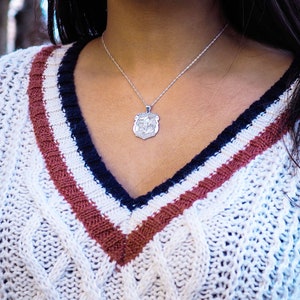 K&ouml;nnte beinhalten: Ein wei&szlig;er Strickpullover mit Zopfmuster und roten und dunkelblauen Streifen. Der Pullover hat einen V-Ausschnitt und eine silberne Kettenkette mit einem Anh&auml;nger.