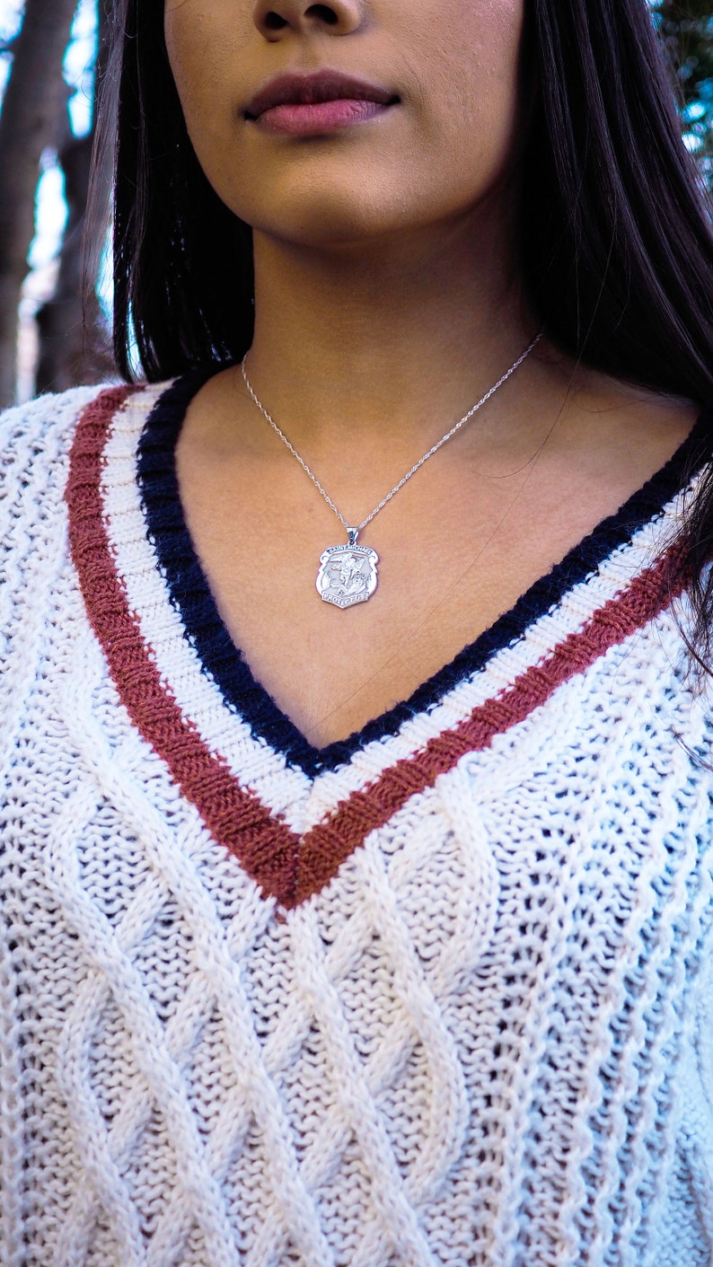 K&ouml;nnte beinhalten: Ein wei&szlig;er Strickpullover mit Zopfmuster und roten und dunkelblauen Streifen. Der Pullover hat einen V-Ausschnitt und eine silberne Kettenkette mit einem Anh&auml;nger.
