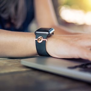 Pu&ograve; includere: Un orologio intelligente nero con un cinturino in oro rosa e un inserto fotografico rotondo con una coppia. L'orologio &egrave; indossato al polso di una persona.