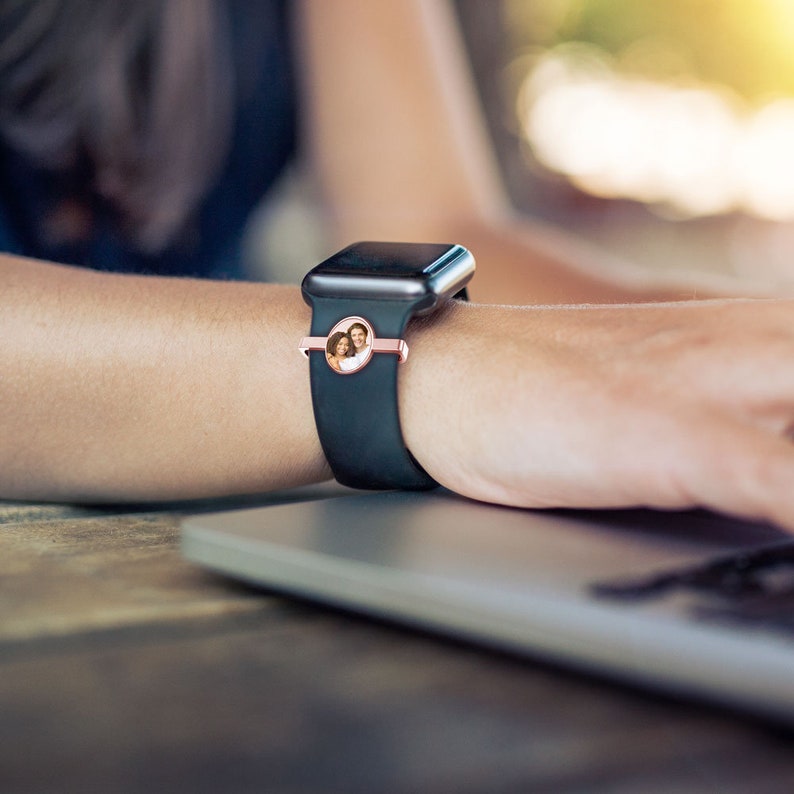Pu&ograve; includere: Un orologio intelligente nero con un cinturino in oro rosa e un inserto fotografico rotondo con una coppia. L'orologio &egrave; indossato al polso di una persona.