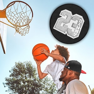 K&ouml;nnte beinhalten: Ein Vater und sein Sohn spielen Basketball. Der Sohn tr&auml;gt ein wei&szlig;es Hemd und h&auml;lt einen Basketball. Der Vater tr&auml;gt eine schwarze Baseballkappe und ein wei&szlig;es Hemd. Der Sohn springt hoch, um den Ball in den Korb zu werfen. Der Korb ist im Hintergrund. Der Vater h&auml;lt die Beine seines Sohnes. Der Sohn l&auml;chelt. Der Vater schaut seinen Sohn an. Der Basketball hat ein schwarz-wei&szlig;es Design mit der Nummer 22 und dem Namen Danny.