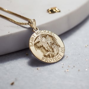 May include: Gold pendant necklace with a round charm featuring an image of Saint Michael the Archangel. The charm has the words "Saint Michael Protect Us" inscribed around the edge.